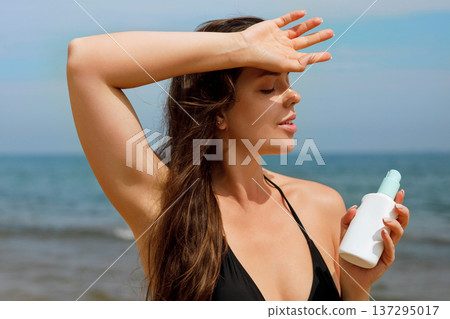 Attractive woman applying sunscreen on face holding suncream lotion bottle at seaside under bright sun Attractive woman applying sunscreen on face holding suncream lotion bottle at seaside under bright sun 137295017