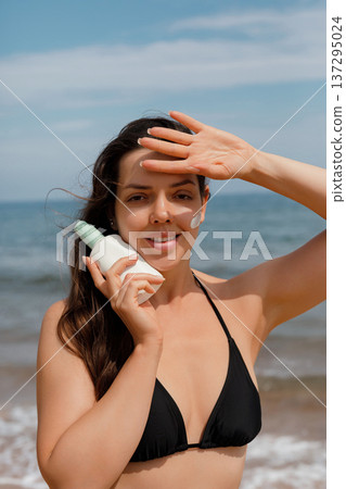 Happy woman shielding eyes from sun while holding sunscreen lotion at beach, summer protection concept Happy woman shielding eyes from sun while holding sunscreen lotion at beach, summer protection concept 137295024
