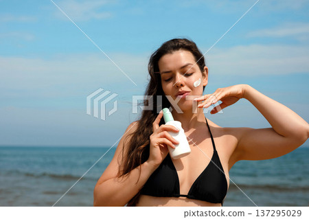 Close portrait of woman applying sunscreen suncream lotion on cheek at beach, female using sunblock for skin care and UV protection Close portrait of woman applying sunscreen suncream lotion on cheek at beach, female using sunblock for skin care and UV protection 137295029