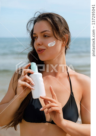 Beautiful woman with sunscreen suncream lotion on cheek holding bottle at beach, female using sunblock for skincare 137295041