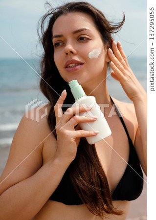 Beautiful woman holding sunscreen suncream lotion and touching face at beach, female using sunblock for skincare routine Beautiful woman holding sunscreen suncream lotion and touching face at beach, female using sunblock for skincare routine 137295056