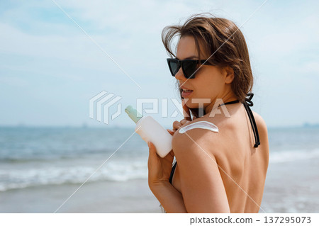 Young woman applying sunscreen lotion on shoulder at beach under sunlight, summer skincare routine and sun protection concept with natural beauty and healthy glowing skin Young woman applying sunscreen lotion on shoulder at beach under sunlight, summer skincare routine and sun protection concept with natural beauty and healthy glowing skin 137295073