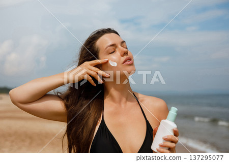 Young woman touching skin while holding sunscreen bottle at seaside, beauty skincare and sun protection concept with healthy glowing face 137295077