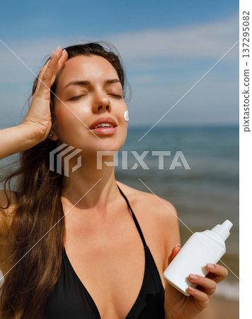 Young woman applying sunscreen lotion on upper body while holding bottle at seaside, summer skincare and sun protection concept with healthy lifestyle Young woman applying sunscreen lotion on upper body while holding bottle at seaside, summer skincare and sun protection concept with healthy lifestyle 137295082