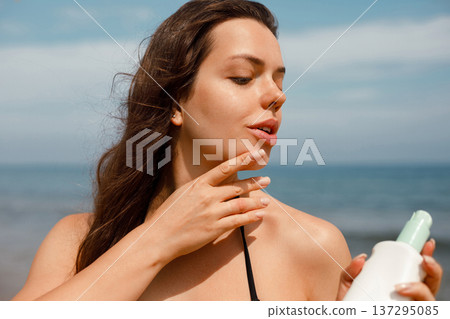 Young woman applying sunscreen lotion on neck holding suncream bottle for UV protection at seaside Young woman applying sunscreen lotion on neck holding suncream bottle for UV protection at seaside 137295085