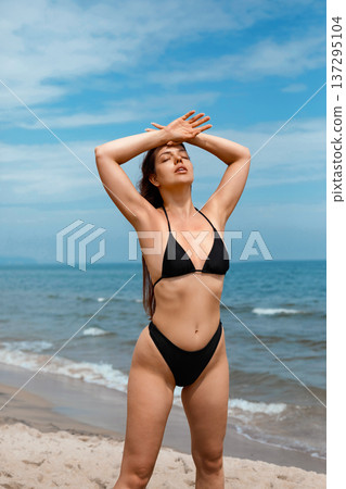 Attractive woman enjoying sunlight on beach in bikini raising arms and using sunscreen suncream lotion for UV protection skincare and summer wellness concept 137295104