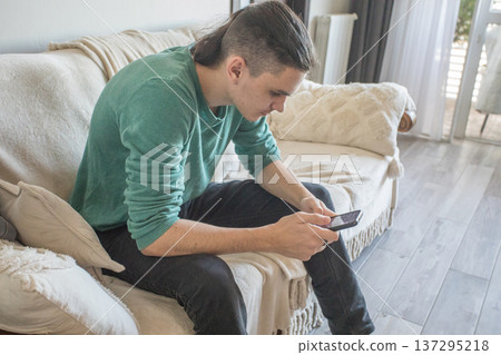 A teenager sits on a couch in a living room playing a video game on a mobile phone 137295218