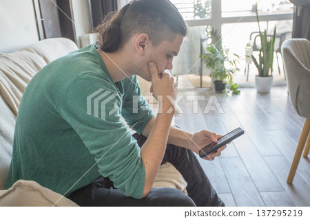 A teenager sits on a couch in a living room playing a video game on a mobile phone 137295219
