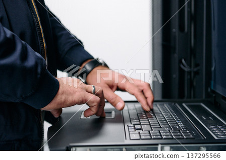 IT technician typing on laptop console in a data center server rack performing maintenance 137295566