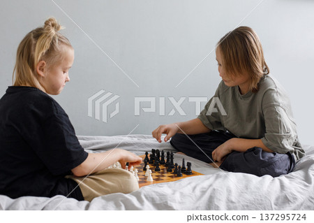 Blond little girl and young boy have a nice game. Sibling playing chess on the bed at home together Blond little girl and young boy have a nice game. Sibling playing chess on the bed at home together 137295724