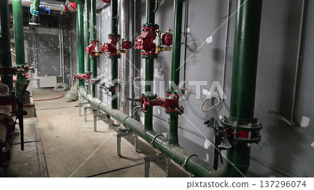 Industrial plumbing setup with green pipes and red valves arranged along a concrete wall in a construction area, showcasing tools and equipment positioned on the floor nearby Industrial plumbing setup with green pipes and red valves arranged along a concrete wall in a construction area, showcasing tools and equipment positioned on the floor nearby 137296074