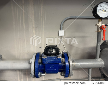Blue water flow meter with a black sensor is attached to gray piping, featuring a pressure gauge on the right side, all mounted on a concrete wall in an industrial environment Blue water flow meter with a black sensor is attached to gray piping, featuring a pressure gauge on the right side, all mounted on a concrete wall in an industrial environment 137296085