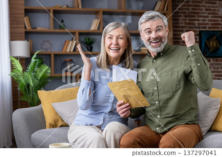 Excited senior couple sitting on a sofa, reading important mail and cheering with happy expressions, celebrating financial success, an investment return, or a retirement announcement at home Excited senior couple sitting on a sofa, reading important mail and cheering with happy expressions, celebrating financial success, an investment return, or a retirement announcement at home 137297041