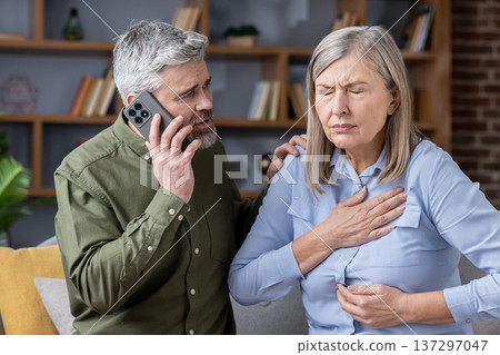 Concerned man dials emergency services while his elderly partner clutches her chest in severe pain at home, urgent medical crisis and support during distress Concerned man dials emergency services while his elderly partner clutches her chest in severe pain at home, urgent medical crisis and support during distress 137297047