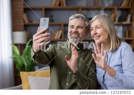 Happy senior couple sitting on a couch, smiling and waving to a handheld smartphone during a video call, communicating with family or friends via technology at home 137297072