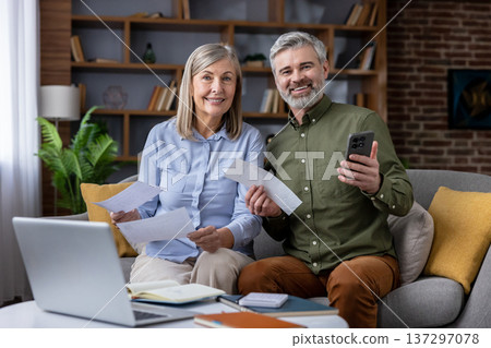 Happy senior couple sitting on a sofa, holding bills and a smartphone while using a laptop to review and manage expenses, representing financial planning and successful budgeting at home Happy senior couple sitting on a sofa, holding bills and a smartphone while using a laptop to review and manage expenses, representing financial planning and successful budgeting at home 137297078