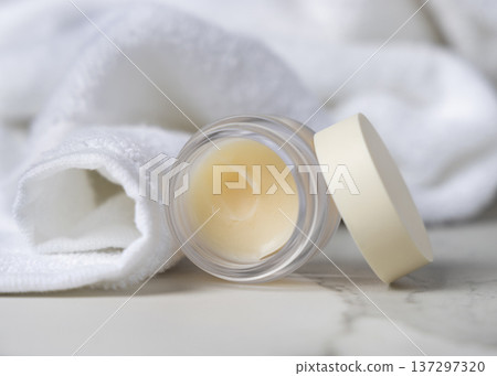 Opened lip balm jar with a lid near white bath towel on stone. Unbranded cosmetic packaging Opened lip balm jar with a lid near white bath towel on stone. Unbranded cosmetic packaging 137297320