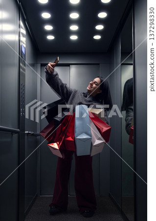Happy woman taking selfie with shopping bags inside modern elevator. 137298330