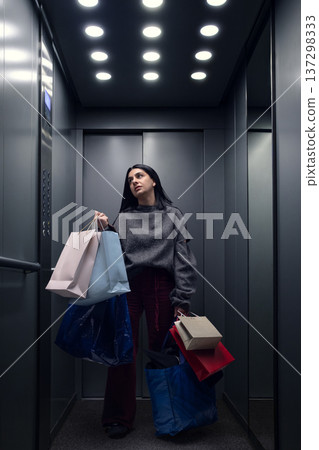 Young woman carrying shopping bags while standing in elevator after retail purchase Young woman carrying shopping bags while standing in elevator after retail purchase 137298333