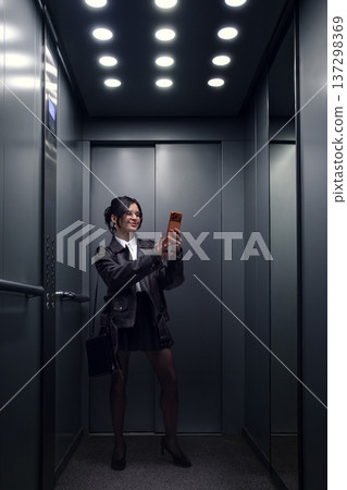 Young woman taking smartphone selfie while standing alone in modern elevator. Young woman taking smartphone selfie while standing alone in modern elevator. 137298369