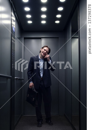 Young businessman smiling while talking on smartphone in modern office elevator. 137298378