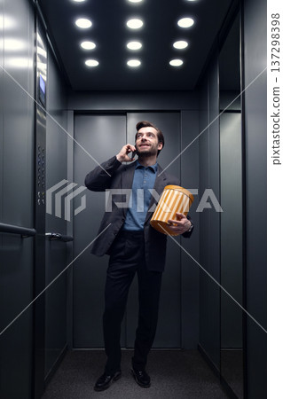 Businessman talking on smartphone while standing in modern office elevator holding gift box. Businessman talking on smartphone while standing in modern office elevator holding gift box. 137298398