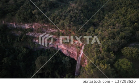 Cassilandia, Mato Grosso do Sul, Brazil - 04 05 2024. Drone aerial view of Cachoeira do Socorro waterfall flowing over a rocky cliff surrounded by dense forest vegetation during early morning light. 137298620