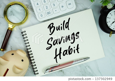Build Savings Habit text written on notepad with piggy bank, calculator, magnifying glass and alarm clock on marble background Build Savings Habit text written on notepad with piggy bank, calculator, magnifying glass and alarm clock on marble background 137302949