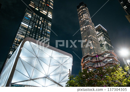 The Vessel sculpture stands illuminated beside modern skyscrapers in Hudson Yards at night. The iconic landmark highlights architectural innovation in New York City. 137304305
