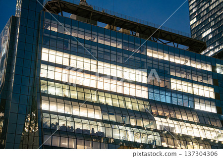 A contemporary glass office building with illuminated floors stands in Hudson Yards at night. The architectural design reflects finance and business activity in New York City. A contemporary glass office building with illuminated floors stands in Hudson Yards at night. The architectural design reflects finance and business activity in New York City. 137304306