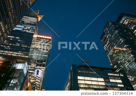 Modern glass skyscrapers glow against the dark night sky in Manhattan. The illuminated towers showcase corporate architecture in New York City. 137304314