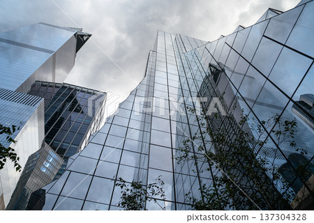 An upward perspective captures glass office towers reflecting the dramatic sky in New York City. The modern skyscrapers symbolize corporate power and urban development. An upward perspective captures glass office towers reflecting the dramatic sky in New York City. The modern skyscrapers symbolize corporate power and urban development. 137304328