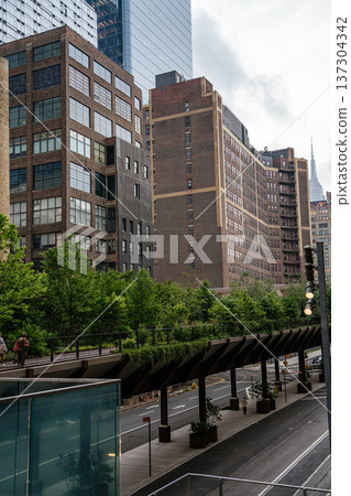 The elevated High Line walkway runs above a Manhattan street lined with historic brick buildings. Modern glass towers rise behind the urban landscape of New York City. The elevated High Line walkway runs above a Manhattan street lined with historic brick buildings. Modern glass towers rise behind the urban landscape of New York City. 137304342