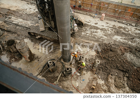 Workers operate drilling equipment while installing a deep foundation pile in a muddy construction area. The scene highlights engineering work, safety gear, and infrastructure development in the city. 137304350