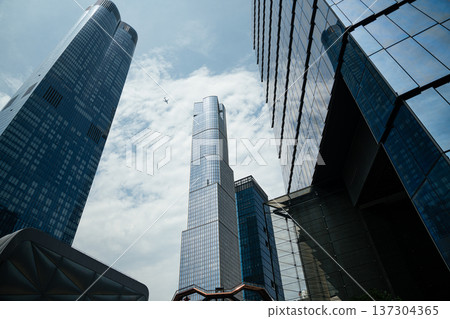 Tall reflective glass skyscrapers rise into a partly cloudy sky in Manhattan with an airplane flying overhead. Contemporary architecture defines the dynamic New York City skyline. Tall reflective glass skyscrapers rise into a partly cloudy sky in Manhattan with an airplane flying overhead. Contemporary architecture defines the dynamic New York City skyline. 137304365