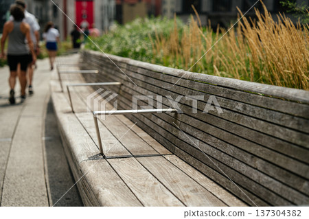 Wooden bench along the High Line elevated park walkway in New York City with ornamental grasses and blurred pedestrians in the background. Urban park design blends nature and modern architecture in Wooden bench along the High Line elevated park walkway in New York City with ornamental grasses and blurred pedestrians in the background. Urban park design blends nature and modern architecture in 137304382
