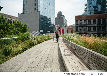 People walking along the High Line elevated park surrounded by greenery and modern buildings in New York City. Urban park pathway offers scenic views of Manhattan architecture and city life. People walking along the High Line elevated park surrounded by greenery and modern buildings in New York City. Urban park pathway offers scenic views of Manhattan architecture and city life. 137304383