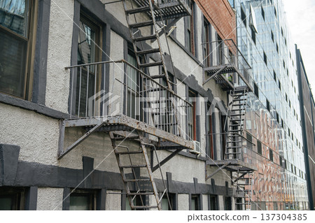 Series of metal fire escapes run along a textured residential facade in Manhattan, New York City. Urban architectural details emphasize safety structures and classic apartment design. Series of metal fire escapes run along a textured residential facade in Manhattan, New York City. Urban architectural details emphasize safety structures and classic apartment design. 137304385