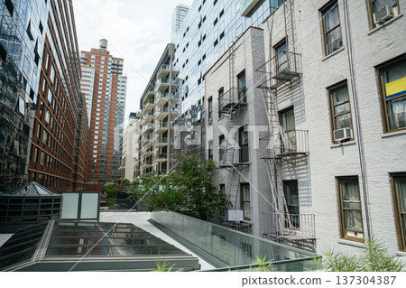 Old metal fire escapes line a white residential building beside modern glass apartments in Manhattan. Contrast between historic and contemporary architecture reflects urban transformation in New York Old metal fire escapes line a white residential building beside modern glass apartments in Manhattan. Contrast between historic and contemporary architecture reflects urban transformation in New York 137304387