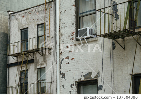 Detailed view of a weathered Manhattan residential wall featuring a metal fire escape and window air conditioner. Textured facade and aging paint reveal classic urban architectural character. 137304396