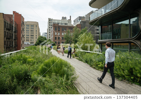 Visitors stroll along the elevated High Line park surrounded by greenery and contemporary architecture in Manhattan. The scene captures urban recreation and modern city design in New York. Visitors stroll along the elevated High Line park surrounded by greenery and contemporary architecture in Manhattan. The scene captures urban recreation and modern city design in New York. 137304405