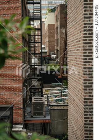 A detailed view of a black metal fire escape rises between tall brick structures in Manhattan. Mechanical rooftop systems and layered facades highlight dense New York architecture. 137304408