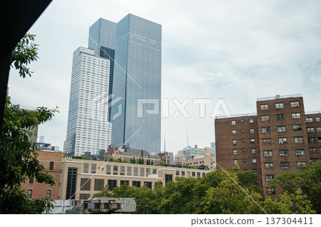 Reflective glass skyscrapers rise beyond brick residential buildings and green trees in Manhattan. The layered skyline shows contrast between historic and modern New York architecture. Reflective glass skyscrapers rise beyond brick residential buildings and green trees in Manhattan. The layered skyline shows contrast between historic and modern New York architecture. 137304411