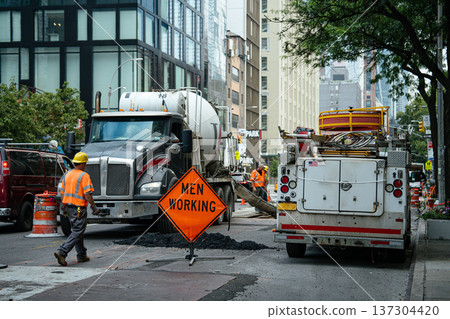Construction workers in safety vests operate near a cement truck on an urban street. A bright orange men working sign warns drivers of active road repairs in the city. Construction workers in safety vests operate near a cement truck on an urban street. A bright orange men working sign warns drivers of active road repairs in the city. 137304420