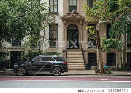 A luxury apartment entrance with decorative details stands behind a parked SUV. Trees and sidewalk frame a refined residential city scene. A luxury apartment entrance with decorative details stands behind a parked SUV. Trees and sidewalk frame a refined residential city scene. 137304424