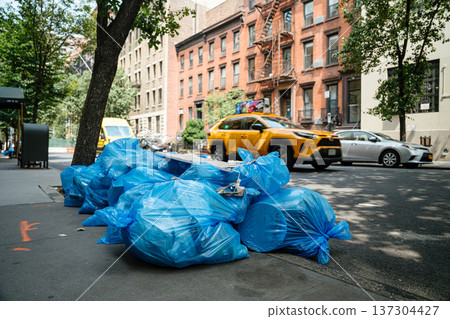 A yellow taxi moves along a tree-lined street beside stacked blue garbage bags on the curb. The image captures everyday transportation, sanitation, and dense urban living. 137304427
