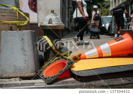 A street fire hydrant is surrounded by fallen orange traffic cones and caution tape on a busy urban sidewalk. Pedestrians walk past construction safety equipment in a downtown city environment. A street fire hydrant is surrounded by fallen orange traffic cones and caution tape on a busy urban sidewalk. Pedestrians walk past construction safety equipment in a downtown city environment. 137304432