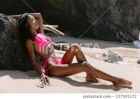 Woman relaxing in pink bikini on sandy beach near rocks, tropical summer vibes Woman relaxing in pink bikini on sandy beach near rocks, tropical summer vibes 137305172