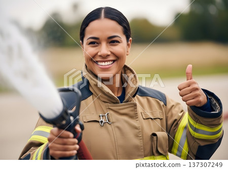 Smiling Female Firefighter Gives Thumbs Up While Spraying Water with a Hose 137307249