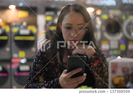 Woman looking at mobile phone while sitting inside modern self-service laundromat Woman looking at mobile phone while sitting inside modern self-service laundromat 137308509
